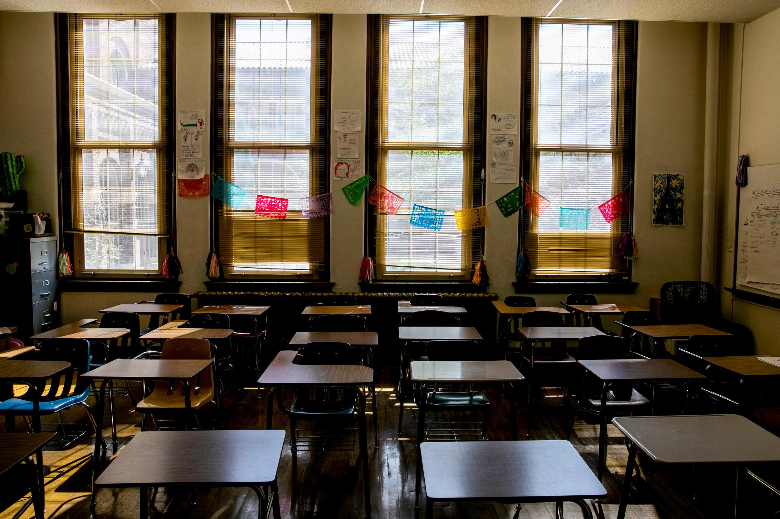 An empty classroom with multiple desks and chairs arranged in rows. The room is lined with windows, providing natural light.