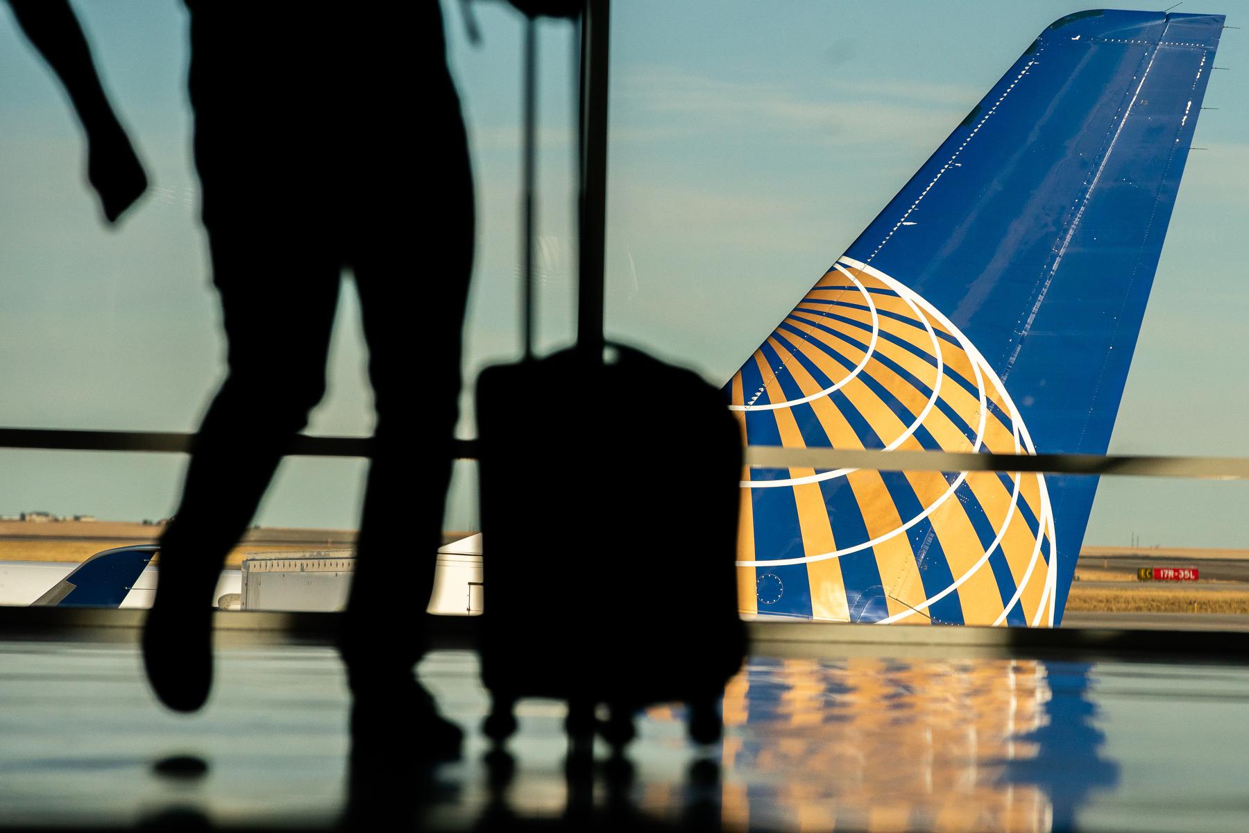The silhouette of a traveler from the waist down with a roller approaches a window showing a view of the tail of a United Airlines plane.