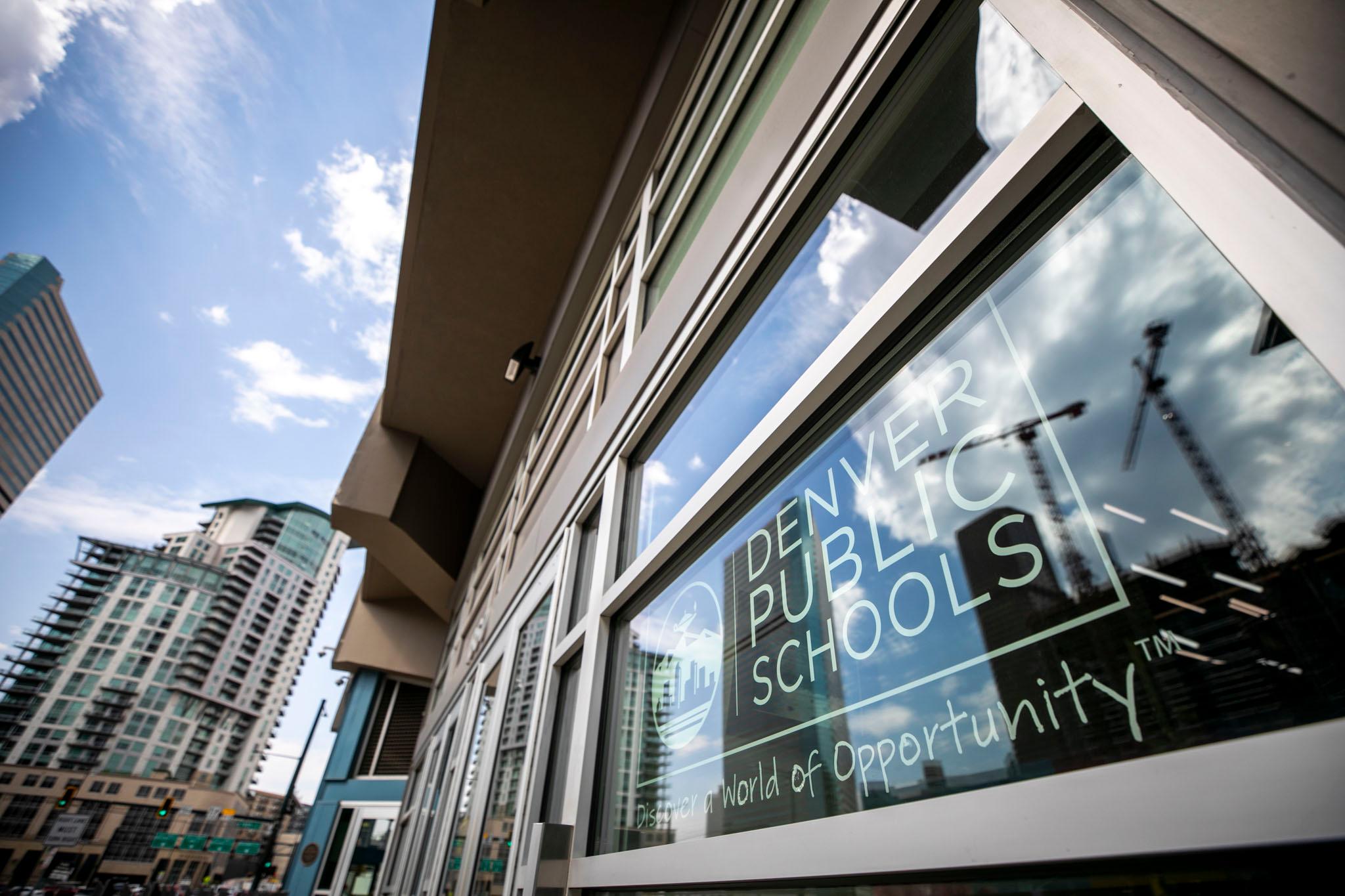 A sign says "Denver Public Schools" in the window of a building; blue skies are reflected in the glass.