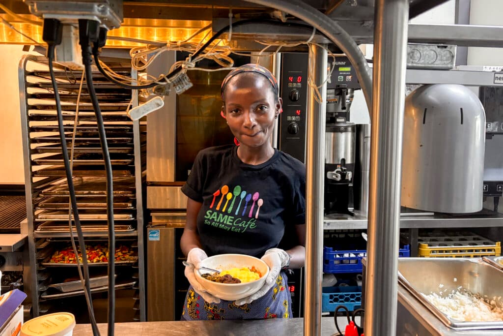 A woman wearing a SAME Café t-shirt and gloves stands in a kitchen, holding a bowl of food and smiling at the camera. She is surrounded by kitchen equipment and trays of prepared food.