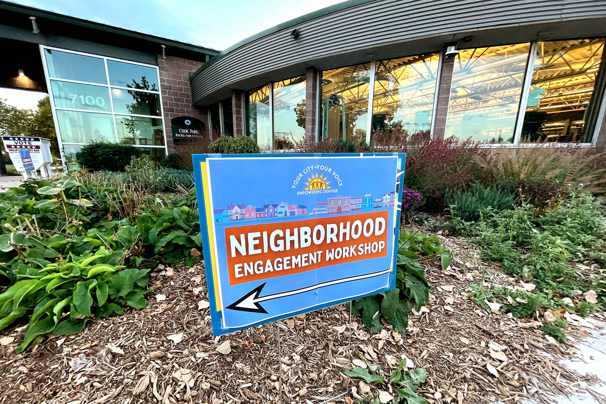 A blue sign with a white arrow is placed in front of a building. It reads: "Neighborhood Engagement Workshop."