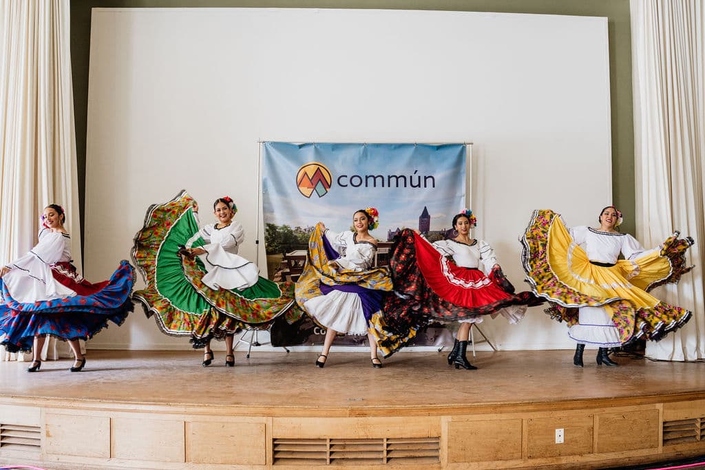 Five dancers in multicolored skirts perform on a wooden stage in front of a Commun banner.