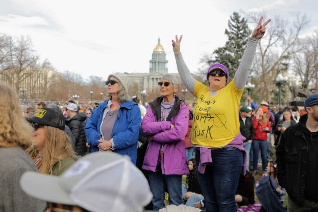 Three women stand in the crowd, wearing colorful light jackets and beanies. One woman's yellow shirt reads &quot;Impeach Trump Deport Musk.&quot;
