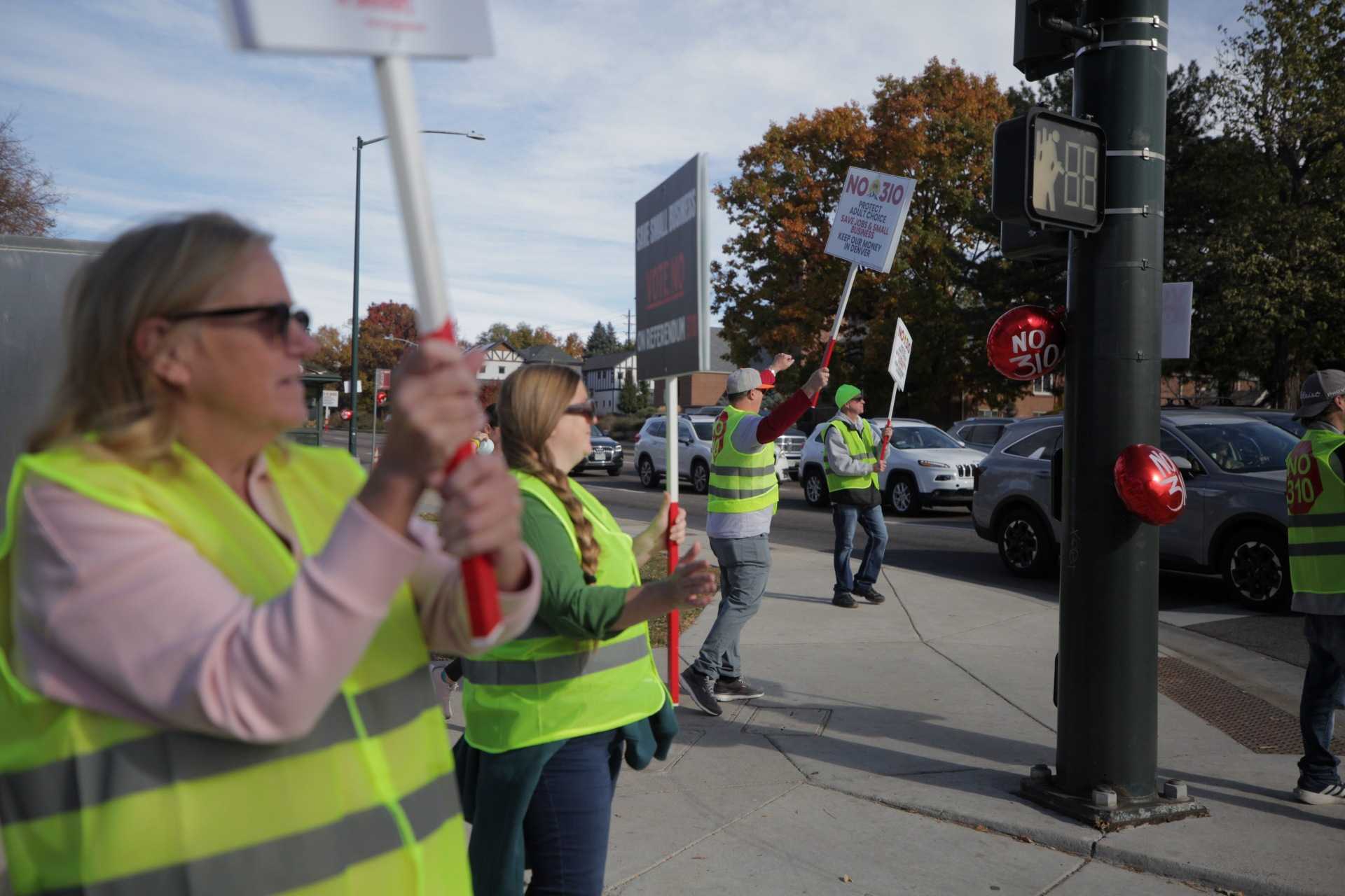People in yellow safety vets hold "No on 310" signs on a busy street corner.