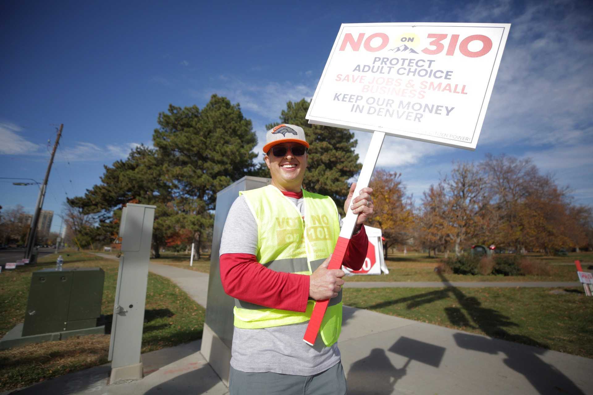 A man, smiling and wearing a yellow safety vest, holds a "No on 310" sign.