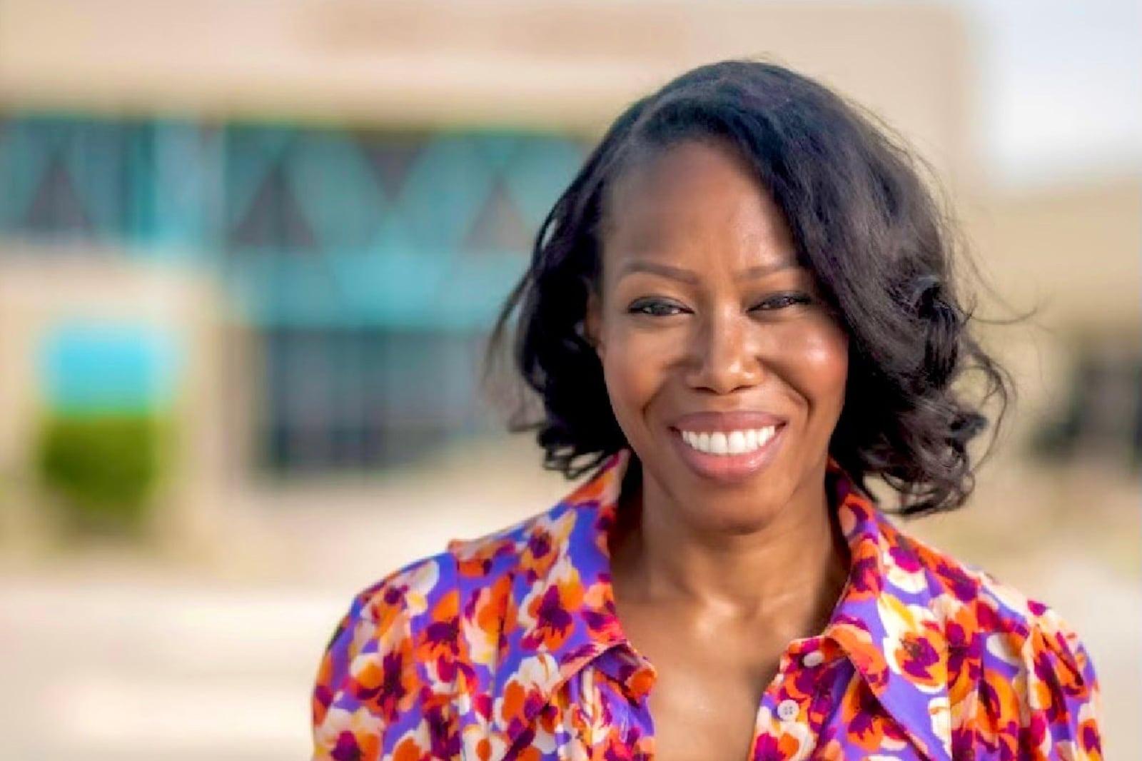 A woman in an orange multi-colored shirt from the collar bone up smiling at the camera with a blury background