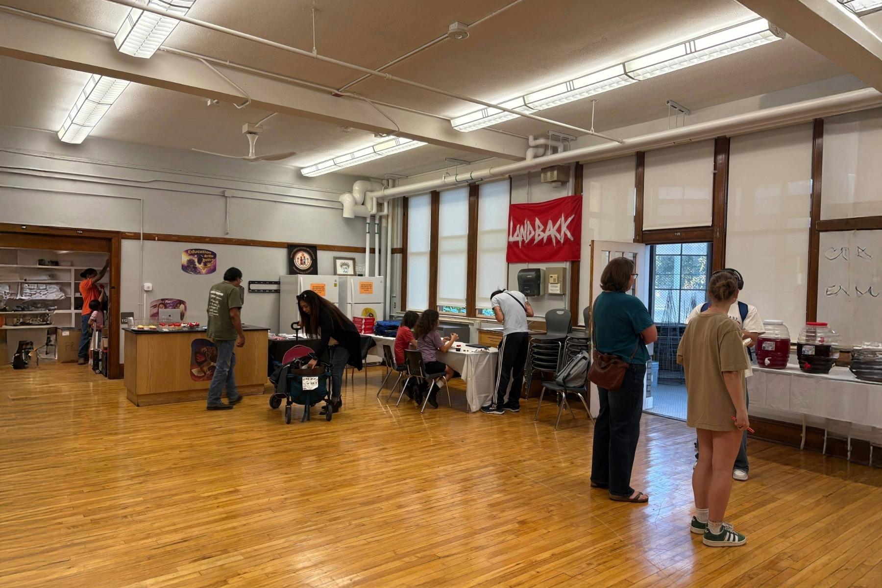 People walk around a large room with wood flooring and fluorescent ceiling lighting. A red sign that says "Land Back" in white letters hangs on the wall.